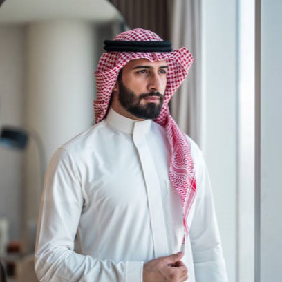 A young Arabic businessman stands confidently by a window, wearing traditional attire with a red and white keffiyeh. The background features modern office elements, adding a professional and focused atmosphere.