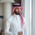 istockphoto-2194497480-612×612 A young Arabic businessman stands confidently by a window, wearing traditional attire with a red and white keffiyeh. The background features modern office elements, adding a professional and focused atmosphere.